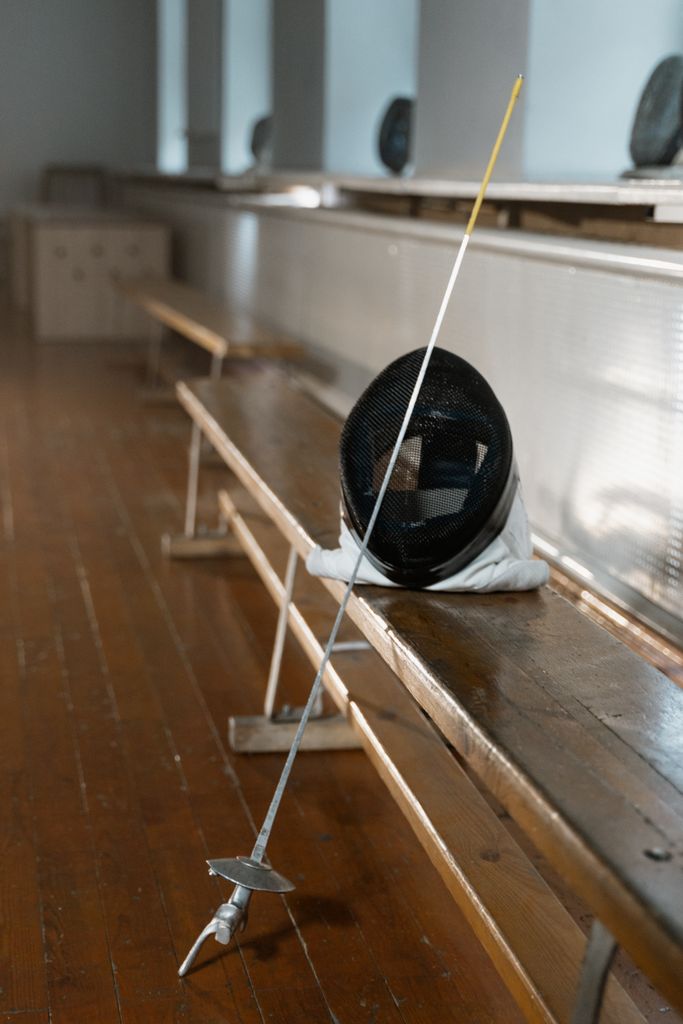 A fencing mask and foil resting on a wooden bench in an indoor arena.